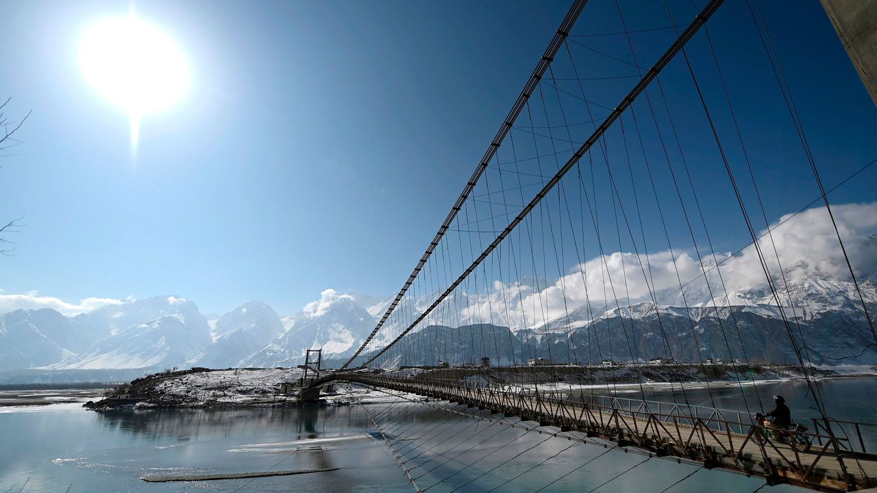 Blue sky, mountainous landscape in the background, with a rope bridge over the Indus River. A motocyclist is crossing the bridge.